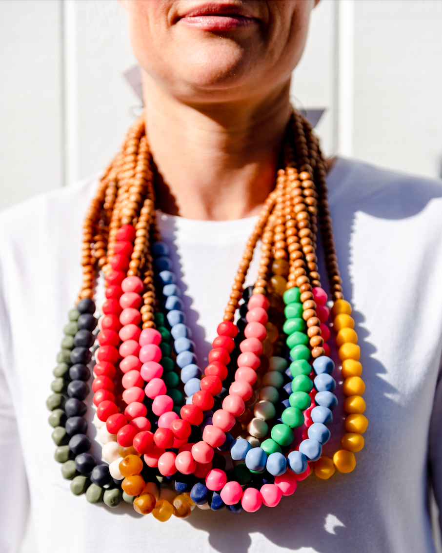 A person wearing a white shirt and showcasing a colorful wooden and rock necklace with multiple strands. A person wearing a white shirt and showcasing a colorful wooden and rock necklace with multiple strands.