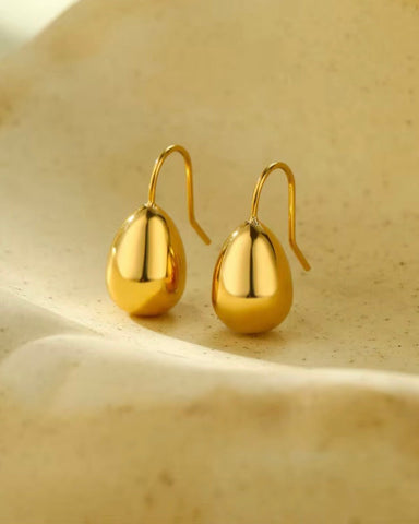 A pair of polished gold teardrop-shaped drop earrings with fishhook backs, displayed on a textured beige stone surface.