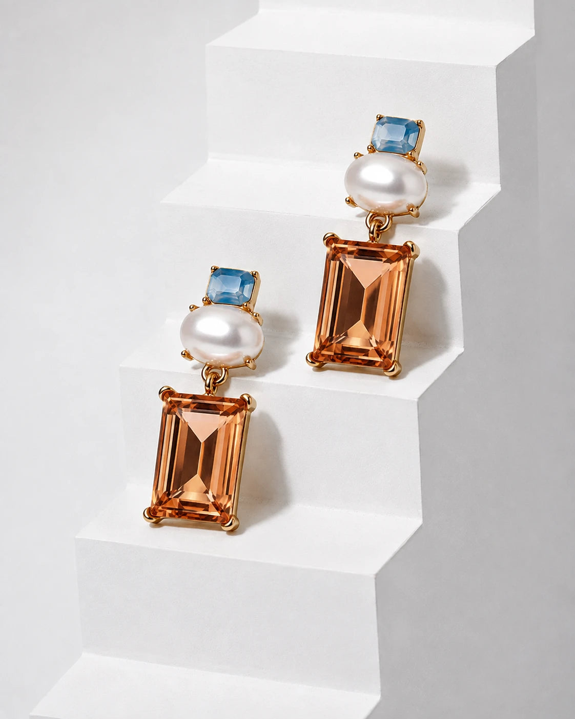 A pair of gold drop earrings with rectangular peach-orange emerald-cut crystals, round white pearls, and small blue square-cut stones, displayed on white geometric steps.