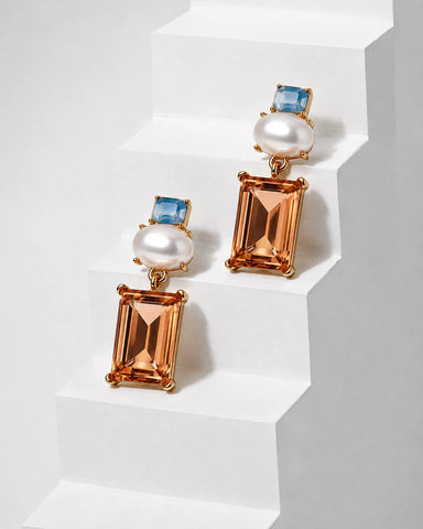 A pair of gold drop earrings with rectangular peach-orange emerald-cut crystals, round white pearls, and small blue square-cut stones, displayed on white geometric steps.