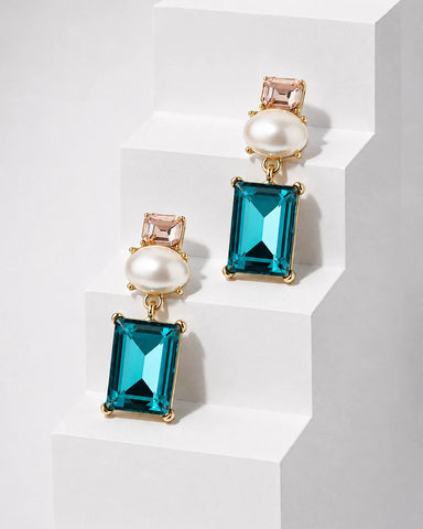A pair of gold-tone drop earrings featuring a large rectangular teal-blue crystal, a white pearl, a small pale pink crystal, and a round pearl, displayed on white geometric steps.