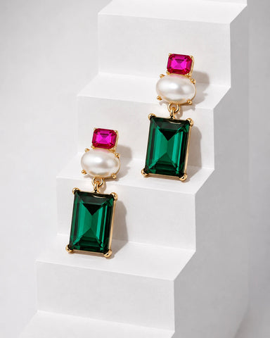 A pair of gold-tone drop earrings featuring a rectangular emerald-cut green crystal, a round white pearl, and a small square pink crystal, displayed on white geometric steps.