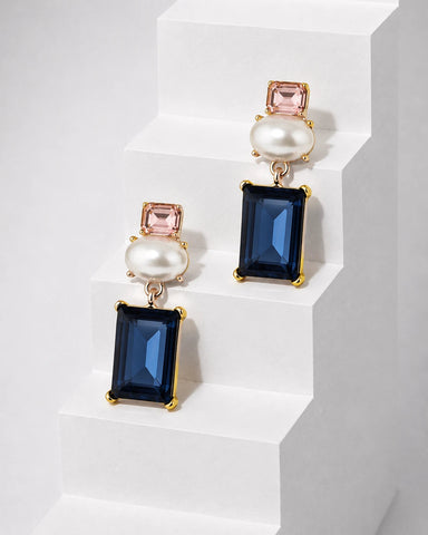 A pair of gold-tone drop earrings with rectangular navy blue gemstones, round white faux pearls, and small pink rectangular gemstones, displayed on white geometric steps.