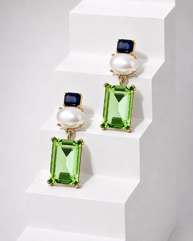 A pair of gold drop earrings featuring rectangular green gemstones, round white pearls, and small dark blue stones, displayed on white geometric steps.