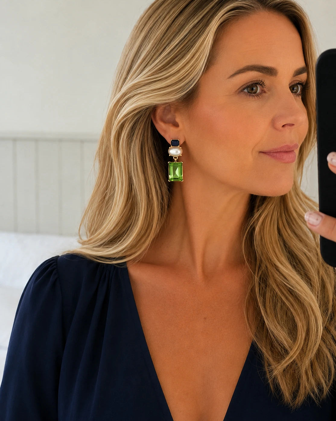 A woman wearing a dangling statement earring with a green rectangular gemstone, a white pearl, and a small dark blue bead, shown in a close-up selfie-style portrait.