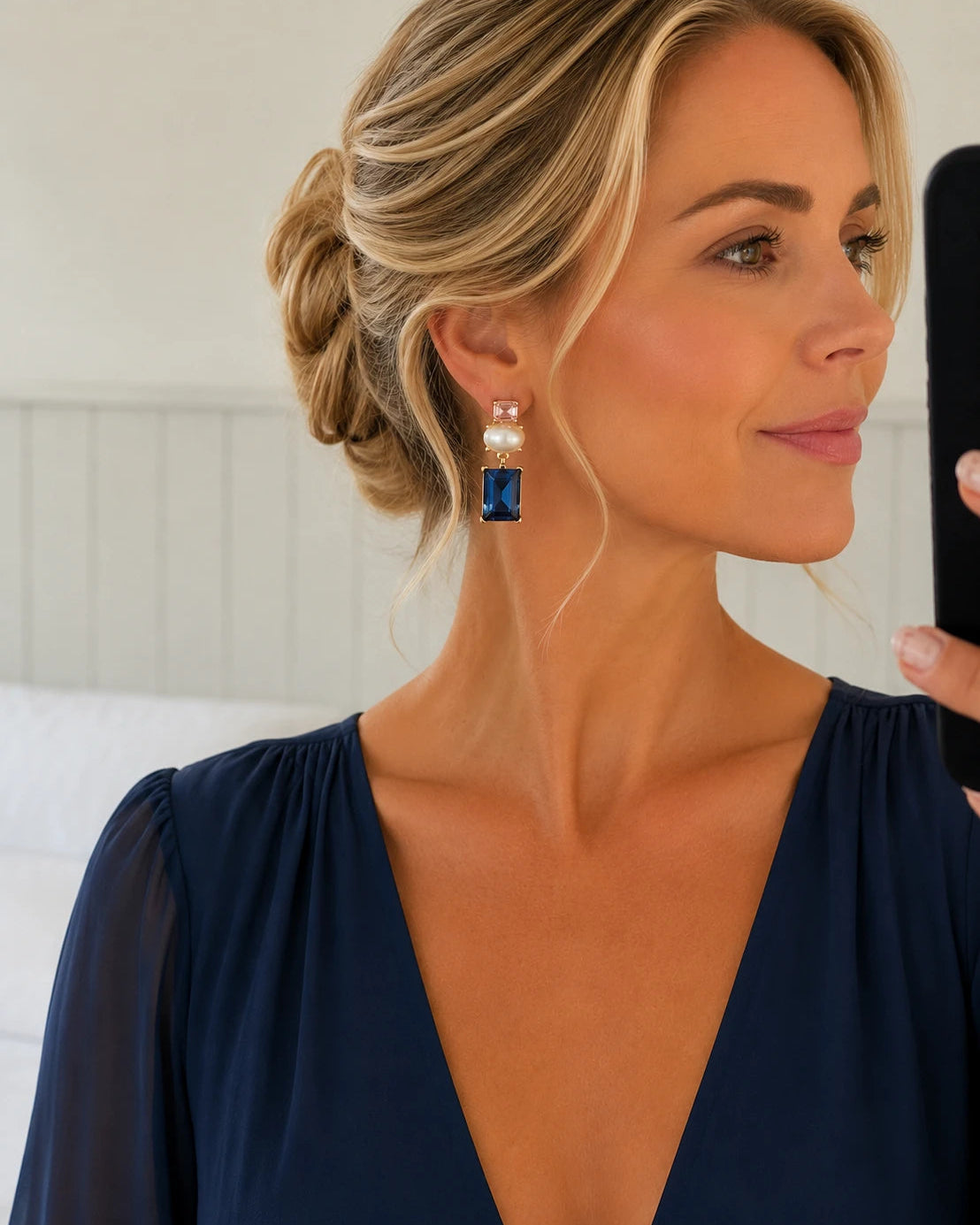 A woman with blonde hair in an updo wearing a dangling earring with a white pearl and a rectangular blue gemstone, holding a phone to take a mirror selfie.