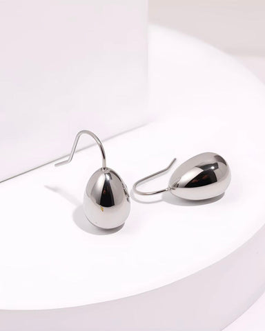 A pair of minimalist silver teardrop-shaped dangle earrings with simple fishhook wires, displayed on a white curved pedestal.