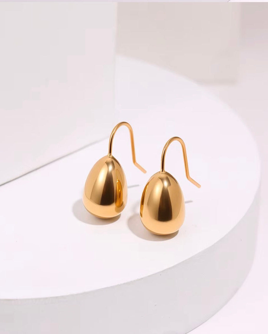 A pair of gold drop earrings with a polished teardrop-shaped drop and a simple hook wire back, displayed on a white curved pedestal.