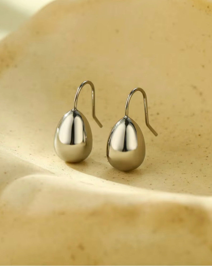 A pair of polished silver teardrop-shaped drop earrings with curved fishhook backs, displayed on a textured beige ceramic dish.