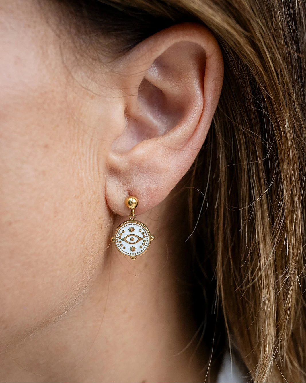 Gold stud earrings with a white enamel evil eye charm dangle