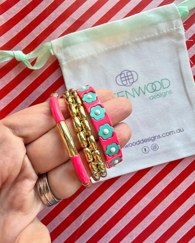 Hand holding three stacked bracelets: a coral pink enamel tube bracelet with a gold bar, a chunky gold link bracelet and a coral pink enamel bracelet with aqua flowers, with a Greenwood Designs calico gift bag.