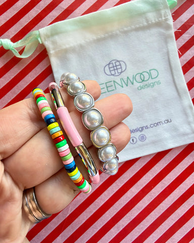 Hand holding three stacked bracelets: a rainbow multicoloured beaded bracelet, a pale pink enamel tube bracelet with a silver bar and a silver-framed pearl bracelet, with a Greenwood Designs calico gift bag.