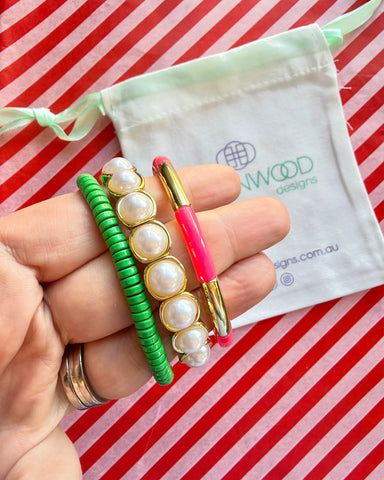 Hand holding three stacked bracelets: a green beaded bracelet, a gold-framed pearl bracelet and a coral pink enamel tube bracelet with a gold bar, with a Greenwood Designs calico gift bag.