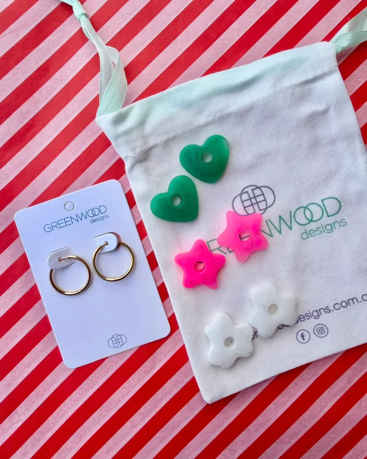 Hoop earrings on a Greenwood Designs card beside a calico pouch topped with three pairs of interchangeable resin charms: green hearts, hot pink stars and white flowers.