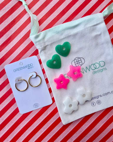 Hoop earrings on a Greenwood Designs card beside a calico pouch topped with three pairs of interchangeable resin charms: green hearts, hot pink stars and white flowers.