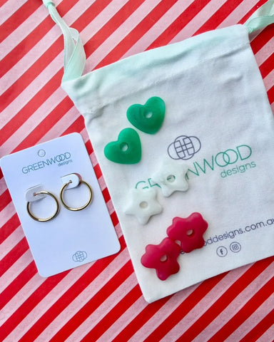 Hoop earrings on a Greenwood Designs card beside a calico pouch topped with three pairs of interchangeable resin charms: green hearts, white stars and raspberry red flowers.