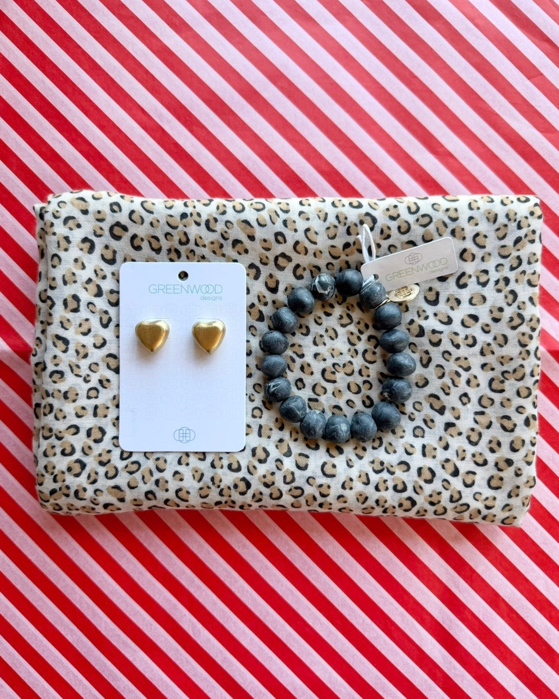 Flat lay of the Sasha leopard print scarf, the Black Marble rock bracelet and Harper Heart earrings in Gold on a Greenwood Designs card, on red and white striped paper.