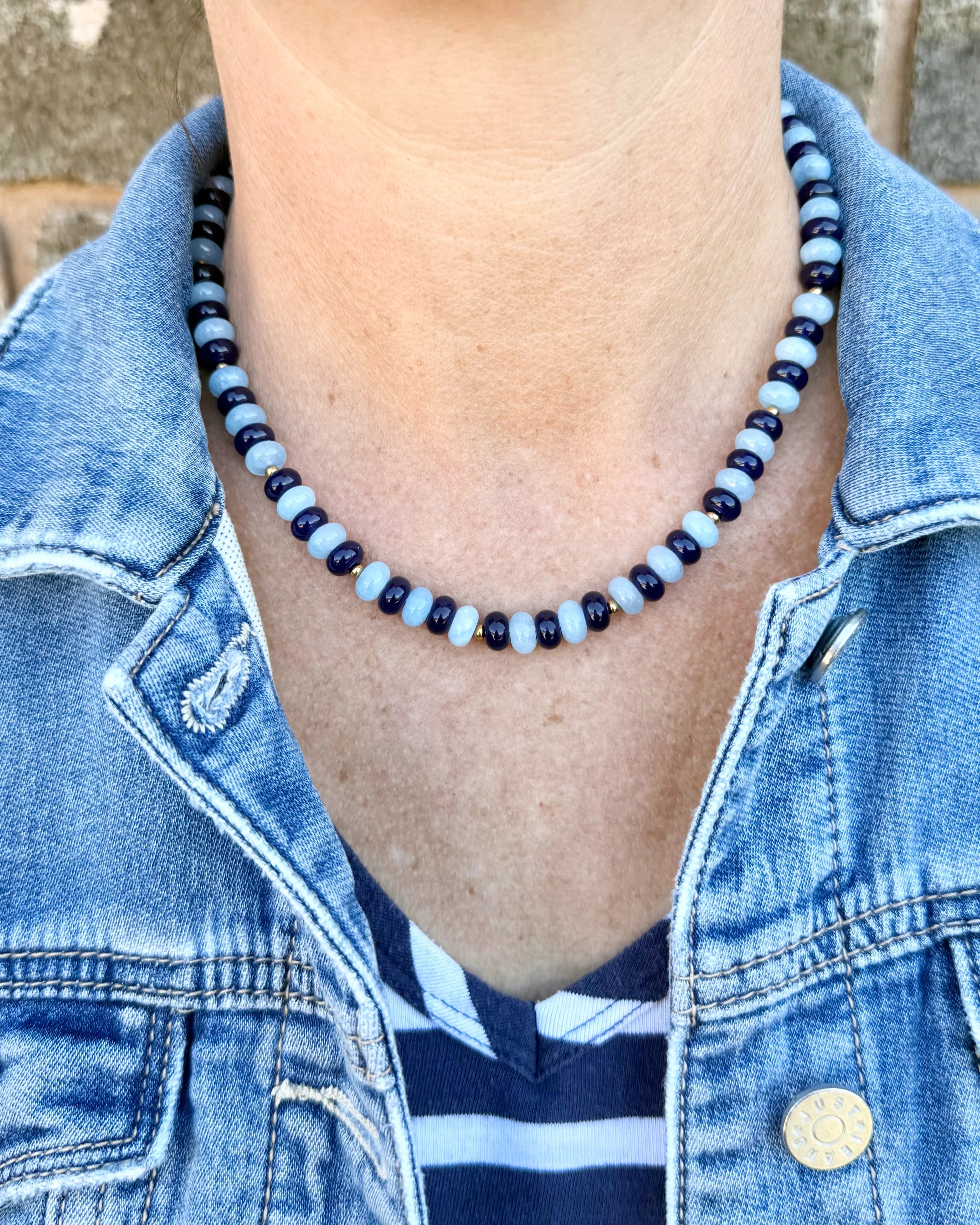 Person wearing a blue denim jacket with a necklace featuring blue and black beads.