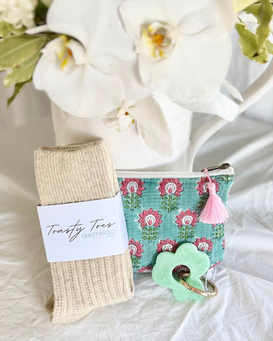 Pair of beige socks with a label, green keychain, and floral pouch on a white surface with flowers in the background.
