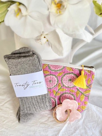 Gray socks with a Toasty Trees label, pink patterned pouch with tassel, and pink keychain on a white surface with white flowers in the background.
