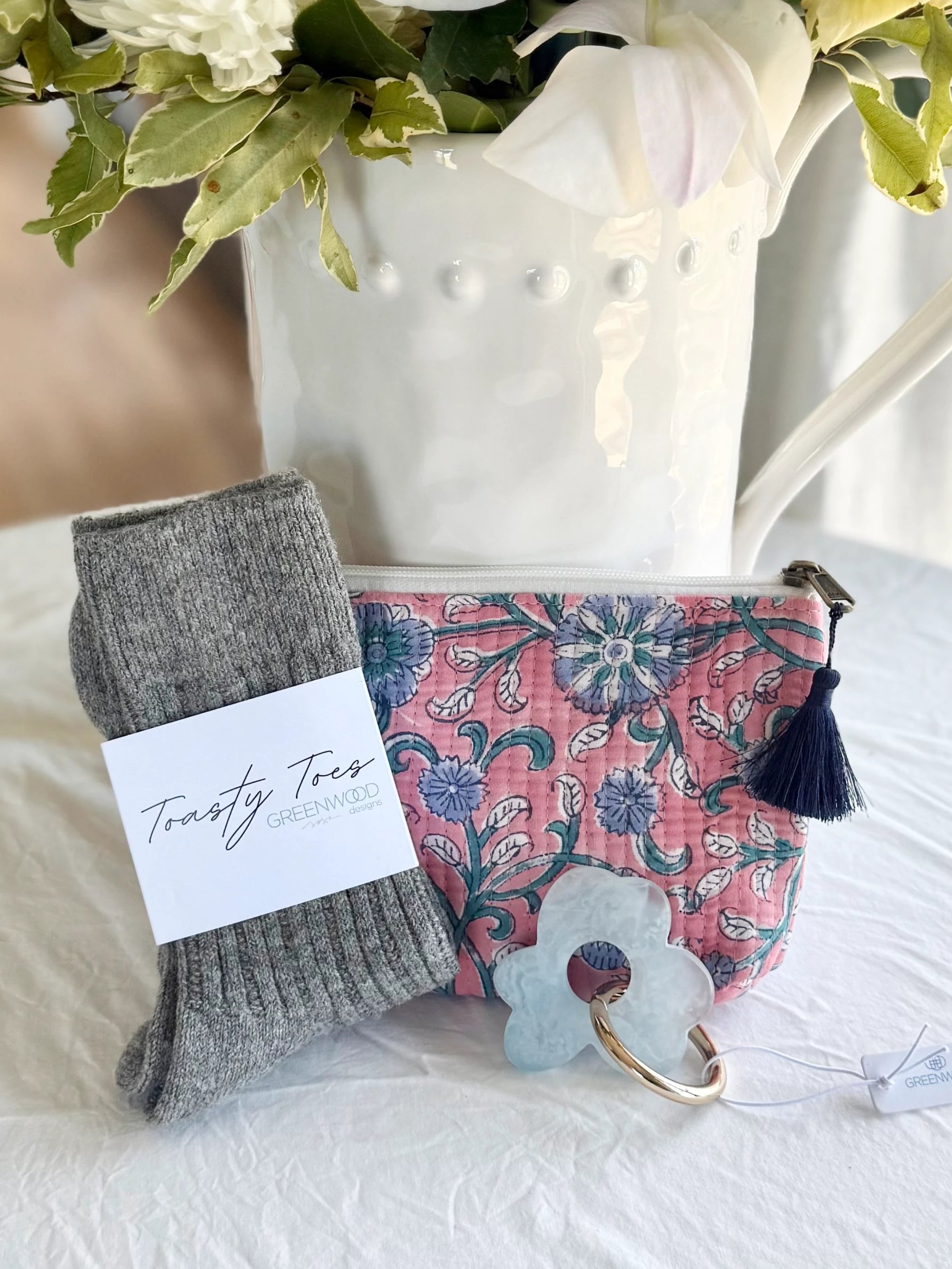Gray socks with a label, pink floral pouch, and keychain on a white surface with flowers in the background.