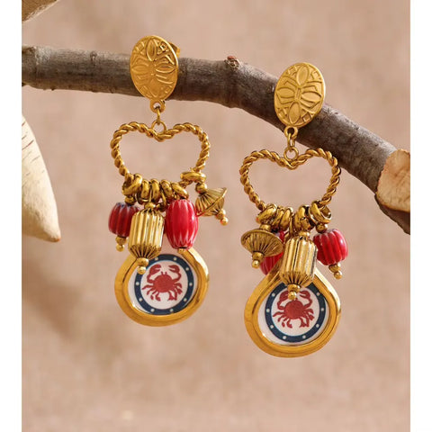 Gold earrings with red striped beads and a gold-framed red crab pendant hanging from a bare branch against a sandy background