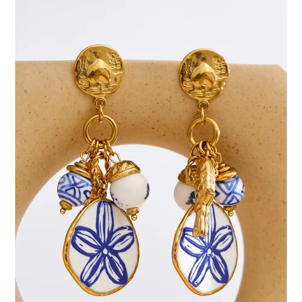 Close-up of gold earrings with blue and white porcelain beads and a white pendant painted with a blue flower, displayed on a beige stand