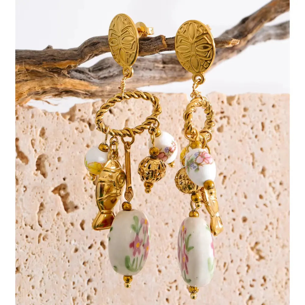 Gold charm earrings with painted floral ceramic beads hanging from a piece of driftwood in front of pale travertine stone