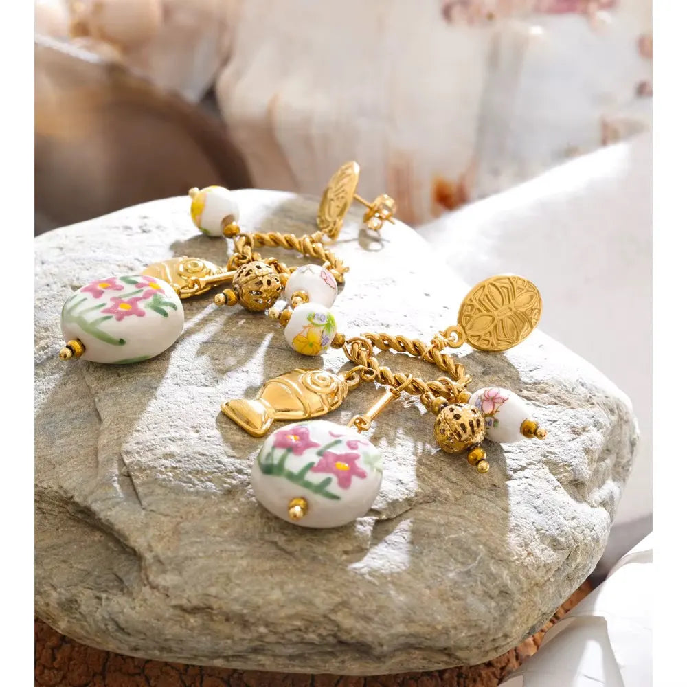 Gold charm earrings with a fish charm and a pink floral ceramic disc resting on a smooth grey stone