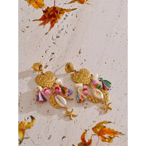 Gold heart tassel earrings with cowrie shells and starfish charms lying on pale stone, scattered with dried orange leaves