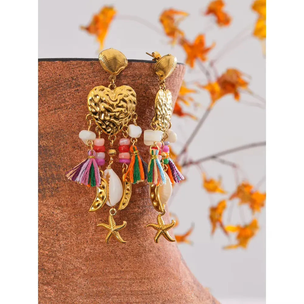 Gold heart earrings with rainbow tassels and cowrie shells hanging from the rim of a terracotta pot, with dried orange flowers behind