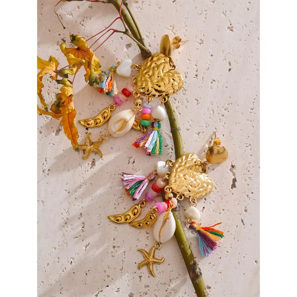 Gold heart earrings with rainbow tassels and cowrie shells hanging from a green stem against pale stone and orange leaves