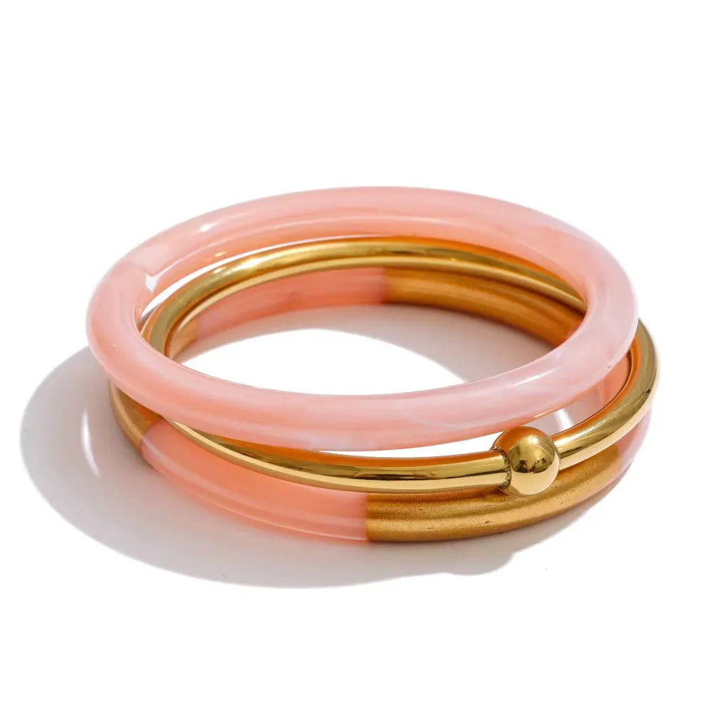 Stack of three bangles: a blush pink resin bangle, a polished gold bangle with a gold ball, and a half pink resin, half gold bangle