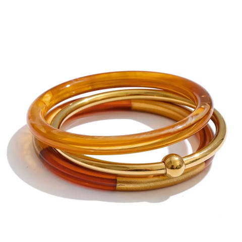 Stack of three bangles: an amber bronze resin bangle, a polished gold bangle with a gold ball, and a half bronze resin, half gold bangle