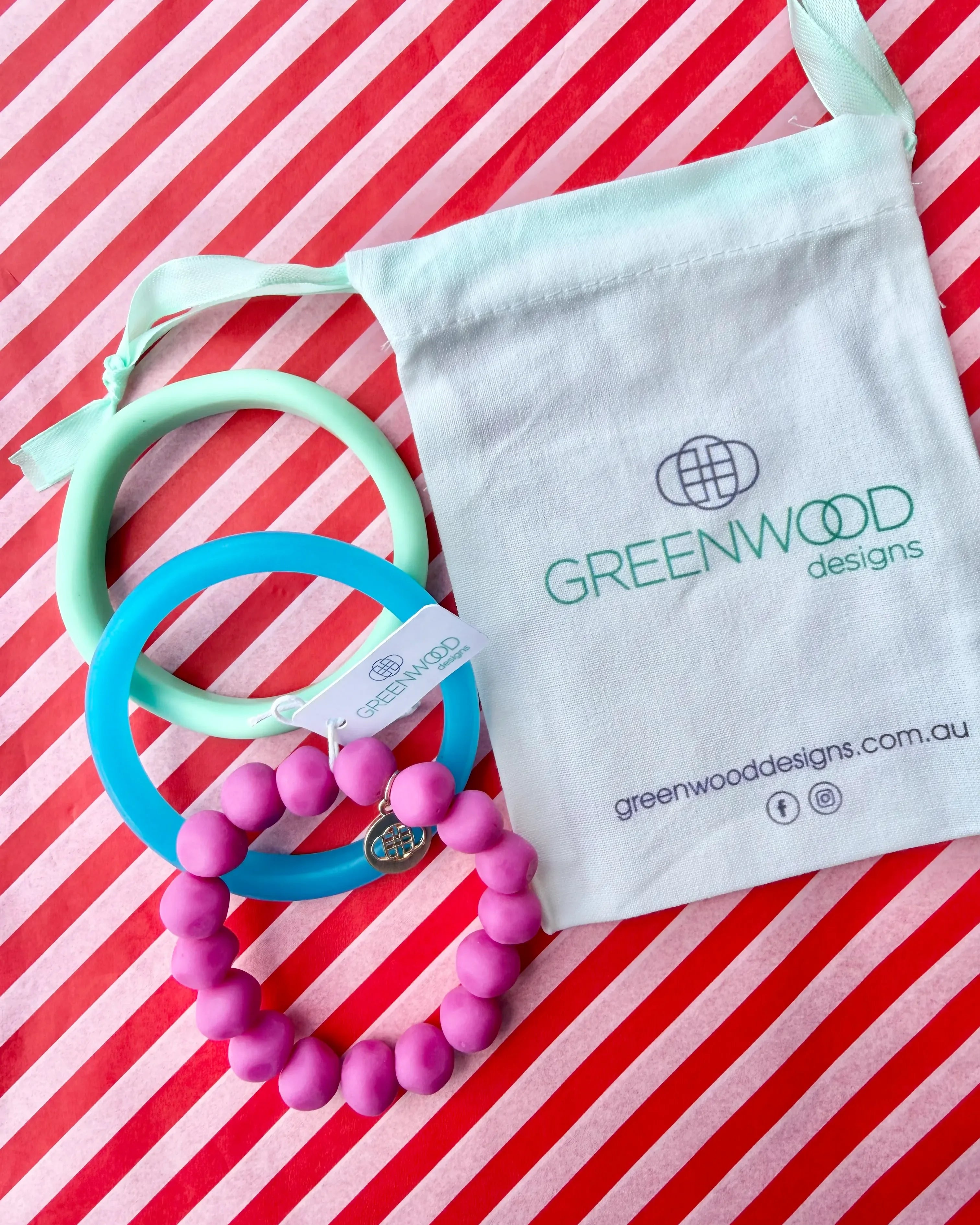An aqua bangle, a sky blue bangle and a fuchsia rock bracelet laid on red and white striped paper beside a Greenwood Designs calico gift bag.