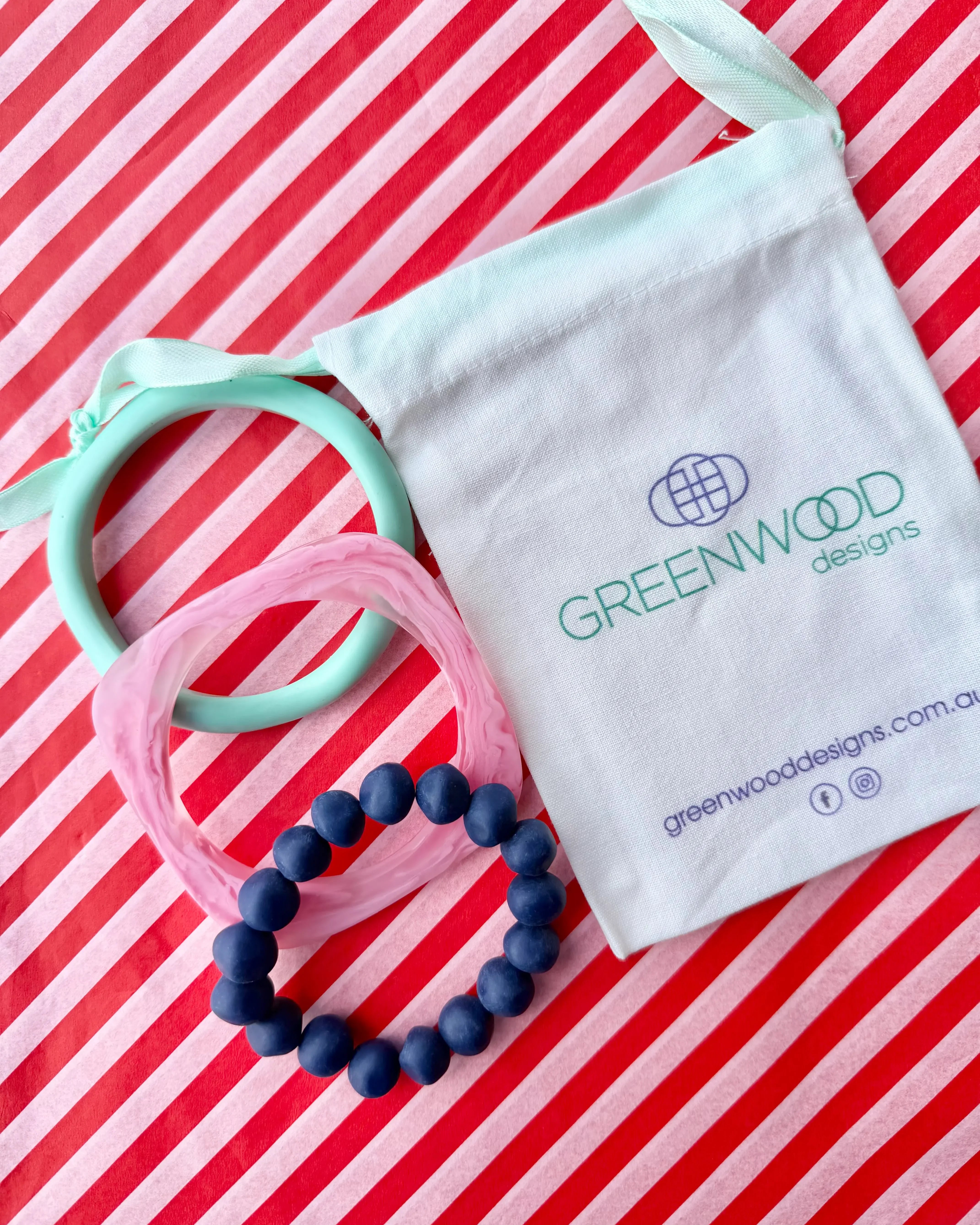An aqua bangle, a light pink marbled geo bangle and a deep blue rock bracelet laid on red and white striped paper beside a Greenwood Designs calico gift bag.
