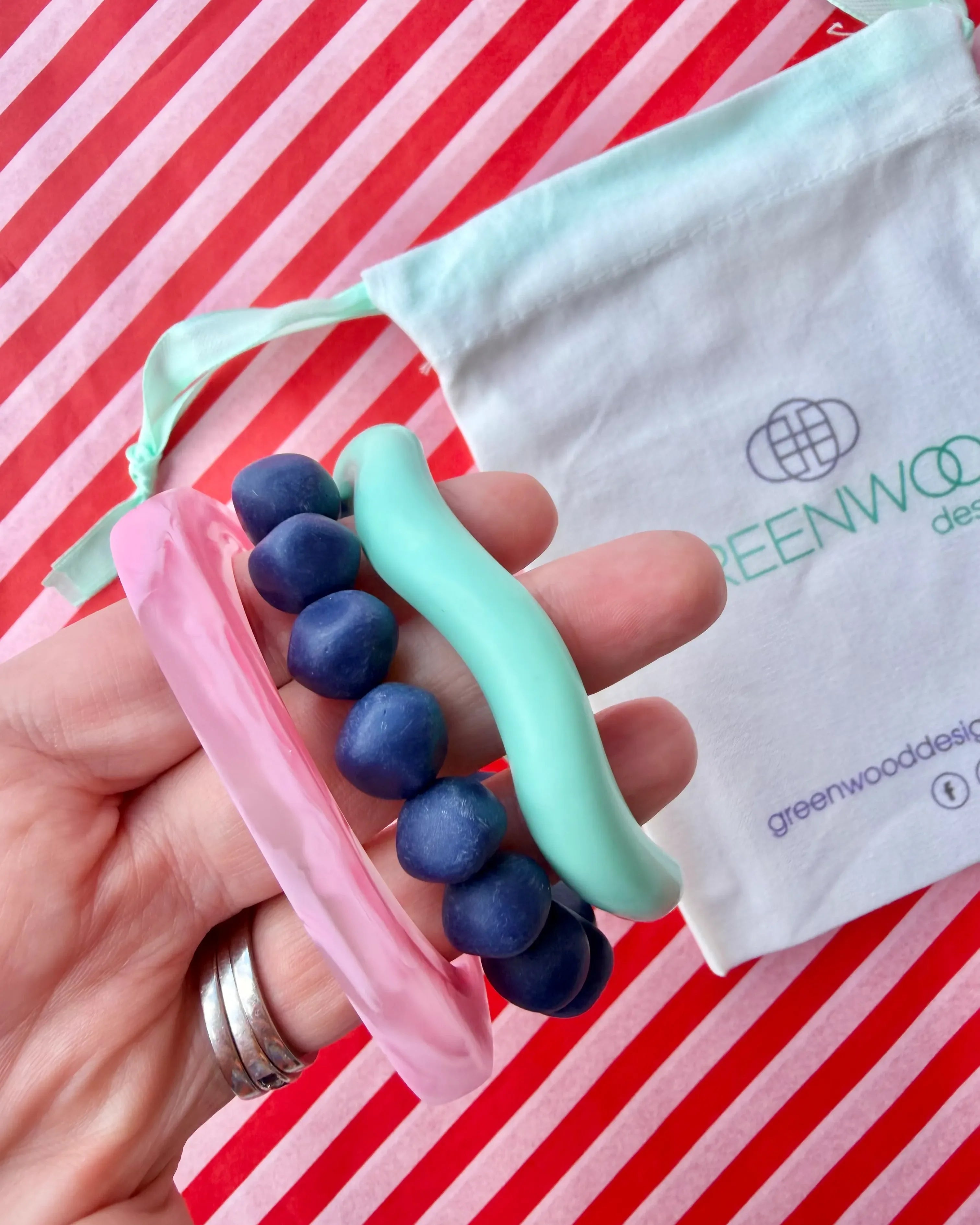 Hand holding a light pink marbled geo bangle, a deep blue rock bracelet and an aqua wave bangle, with a Greenwood Designs calico gift bag on red and white striped paper.