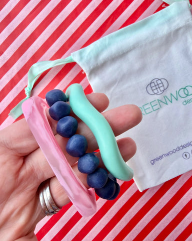Hand holding a light pink marbled geo bangle, a deep blue rock bracelet and an aqua wave bangle, with a Greenwood Designs calico gift bag on red and white striped paper.