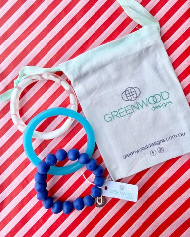 A white marbled wave bangle, a sky blue bangle and a royal blue rock bracelet laid on red and white striped paper beside a Greenwood Designs calico gift bag.