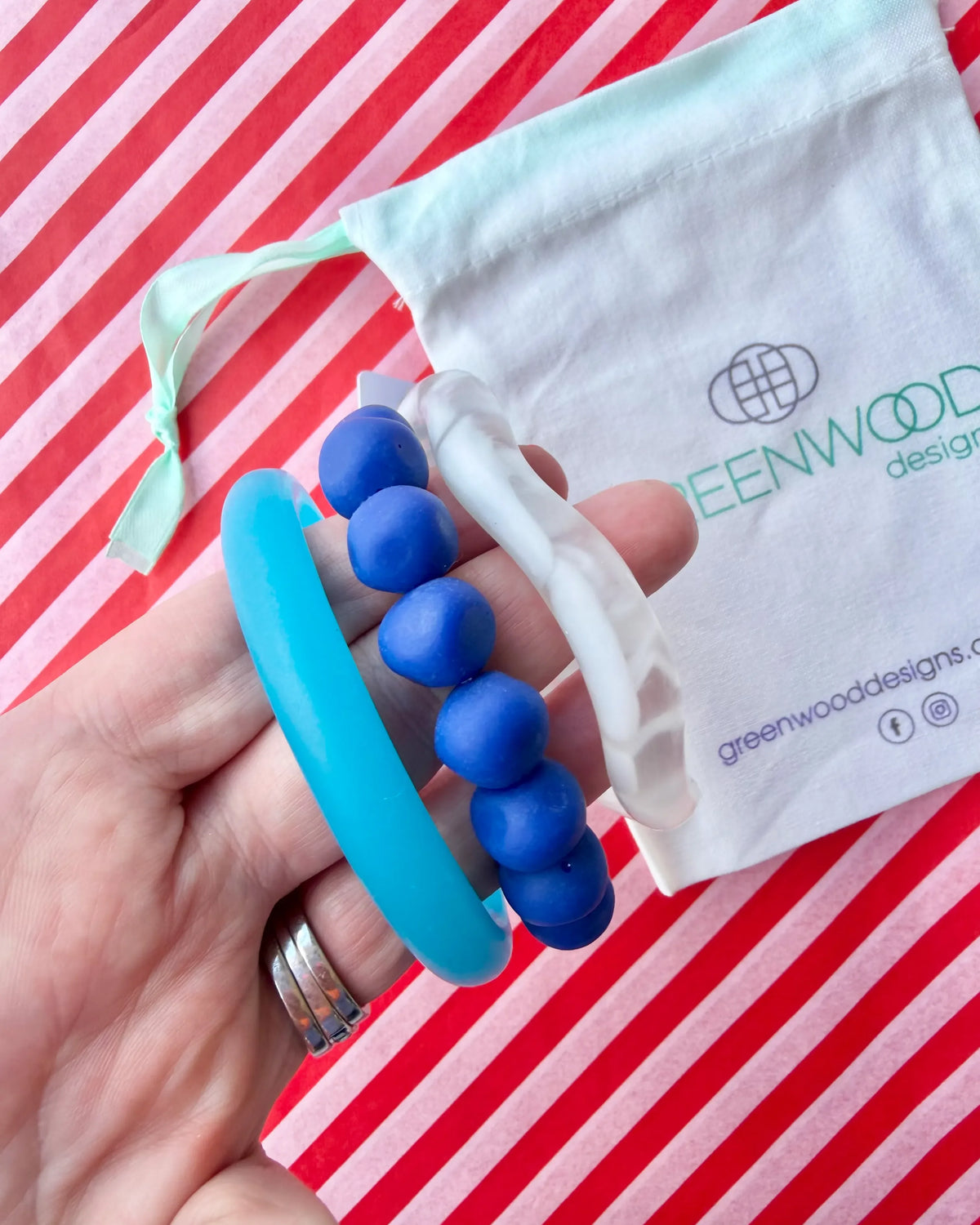 Hand holding a sky blue resin bangle, a royal blue rock bracelet and a white marbled wave bangle, with a Greenwood Designs calico gift bag on red and white striped paper.