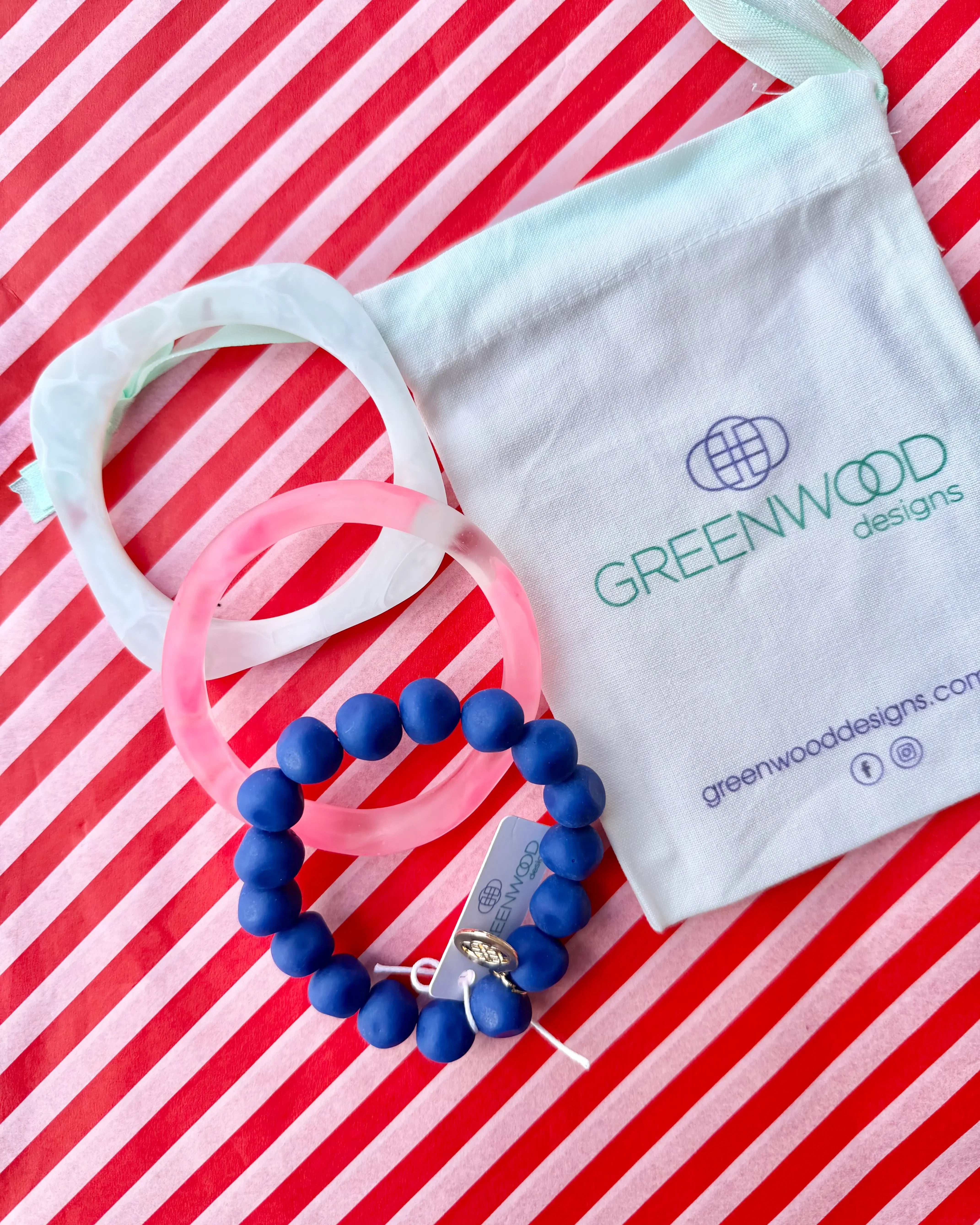 A white marbled geo bangle, a pink marbled wave bangle and a blue rock bracelet laid on red and white striped paper beside a Greenwood Designs calico gift bag.
