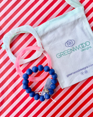 A white marbled geo bangle, a pink marbled wave bangle and a blue rock bracelet laid on red and white striped paper beside a Greenwood Designs calico gift bag.