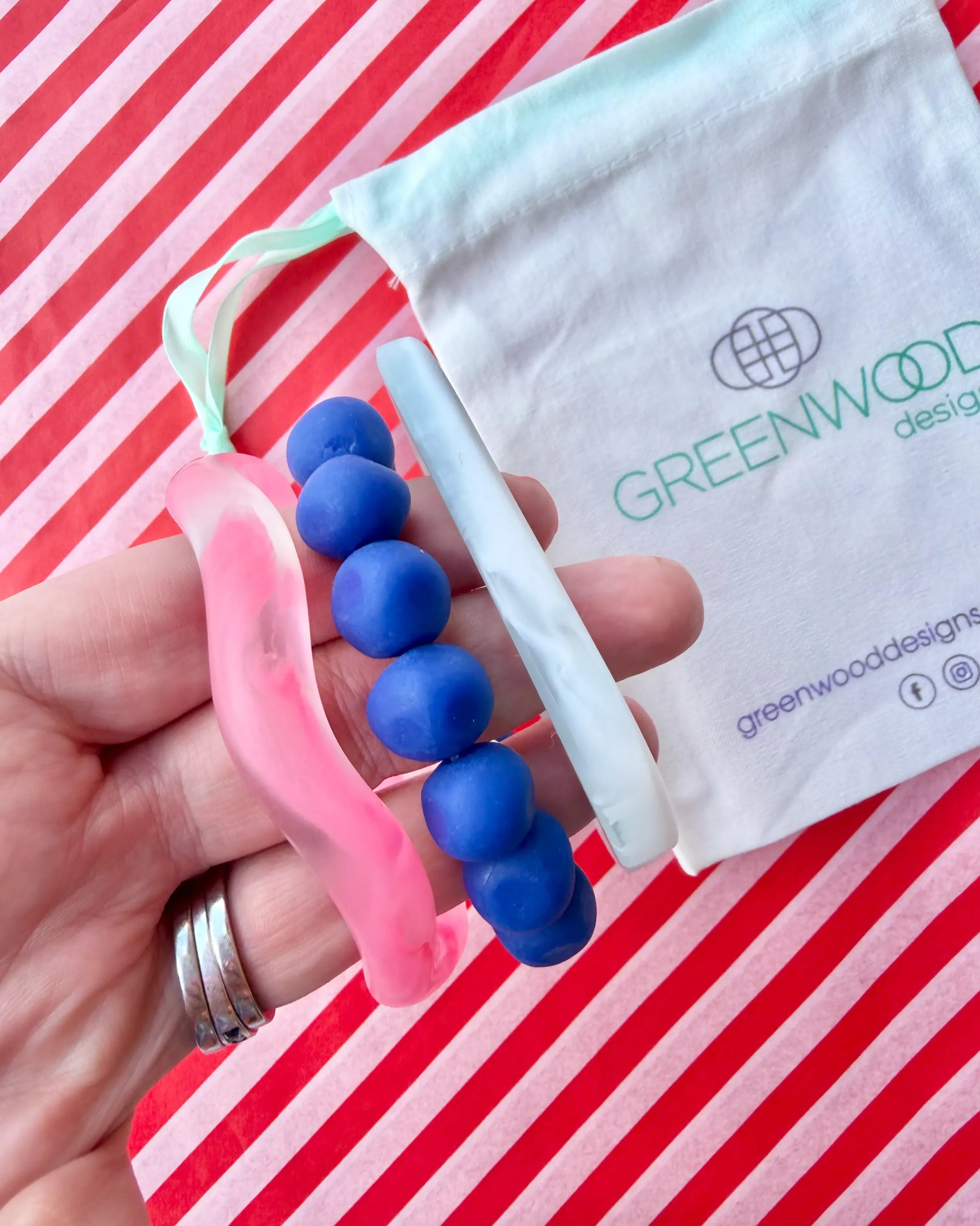 Hand holding a hot pink marbled wave bangle, a blue rock bracelet and a white marbled geo bangle, with a Greenwood Designs calico gift bag on red and white striped paper.