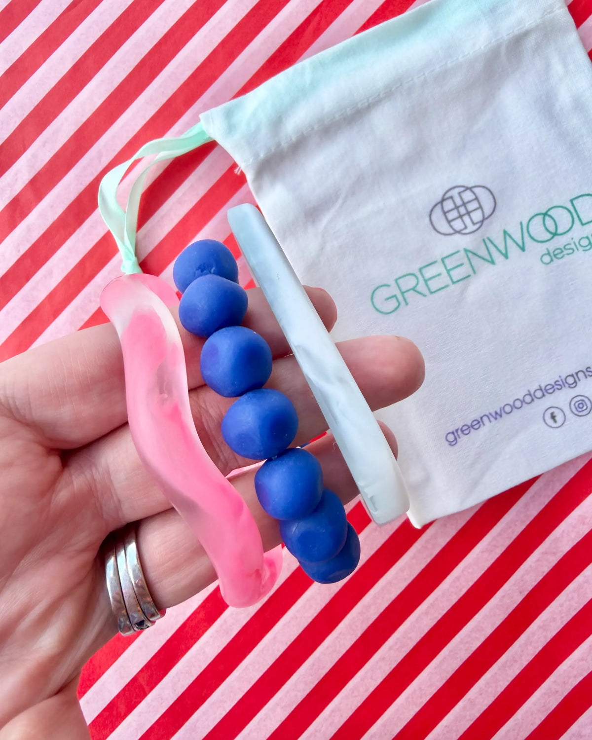 Hand holding a hot pink marbled wave bangle, a blue rock bracelet and a white marbled geo bangle, with a Greenwood Designs calico gift bag on red and white striped paper.