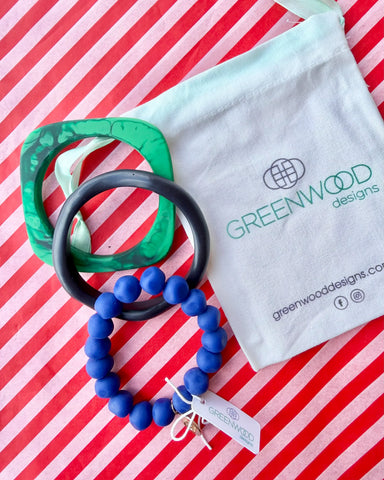 A green marbled geo bangle, a navy bangle and a blue rock bracelet laid on red and white striped paper beside a Greenwood Designs calico gift bag.