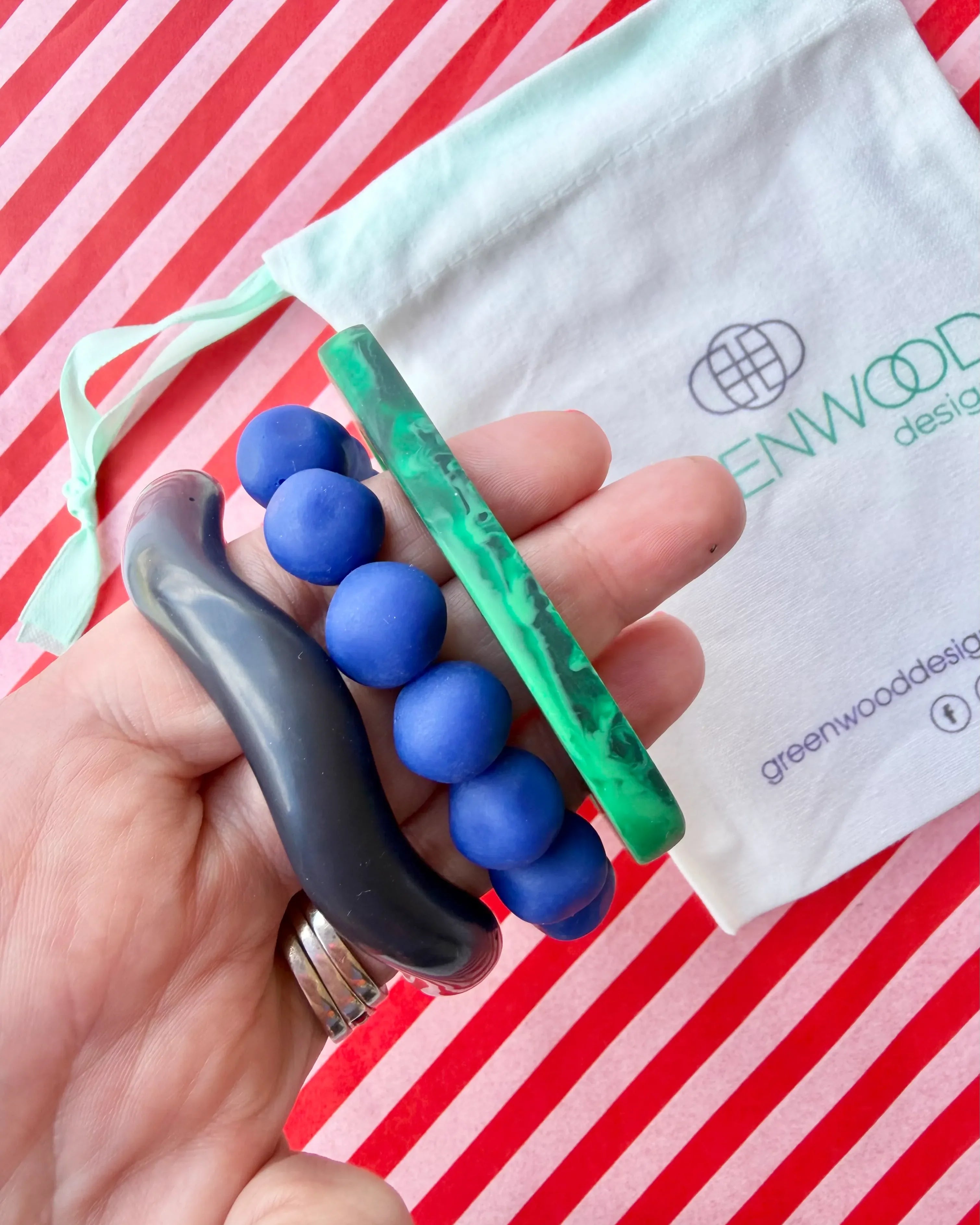 Hand holding a French navy wave bangle, a blue rock bracelet and a green marbled geo bangle, with a Greenwood Designs calico gift bag on red and white striped paper.