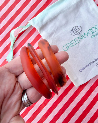 Hand holding three stacked amber tortoiseshell resin bangles in different shapes, with a Greenwood Designs calico gift bag on red and white striped paper.