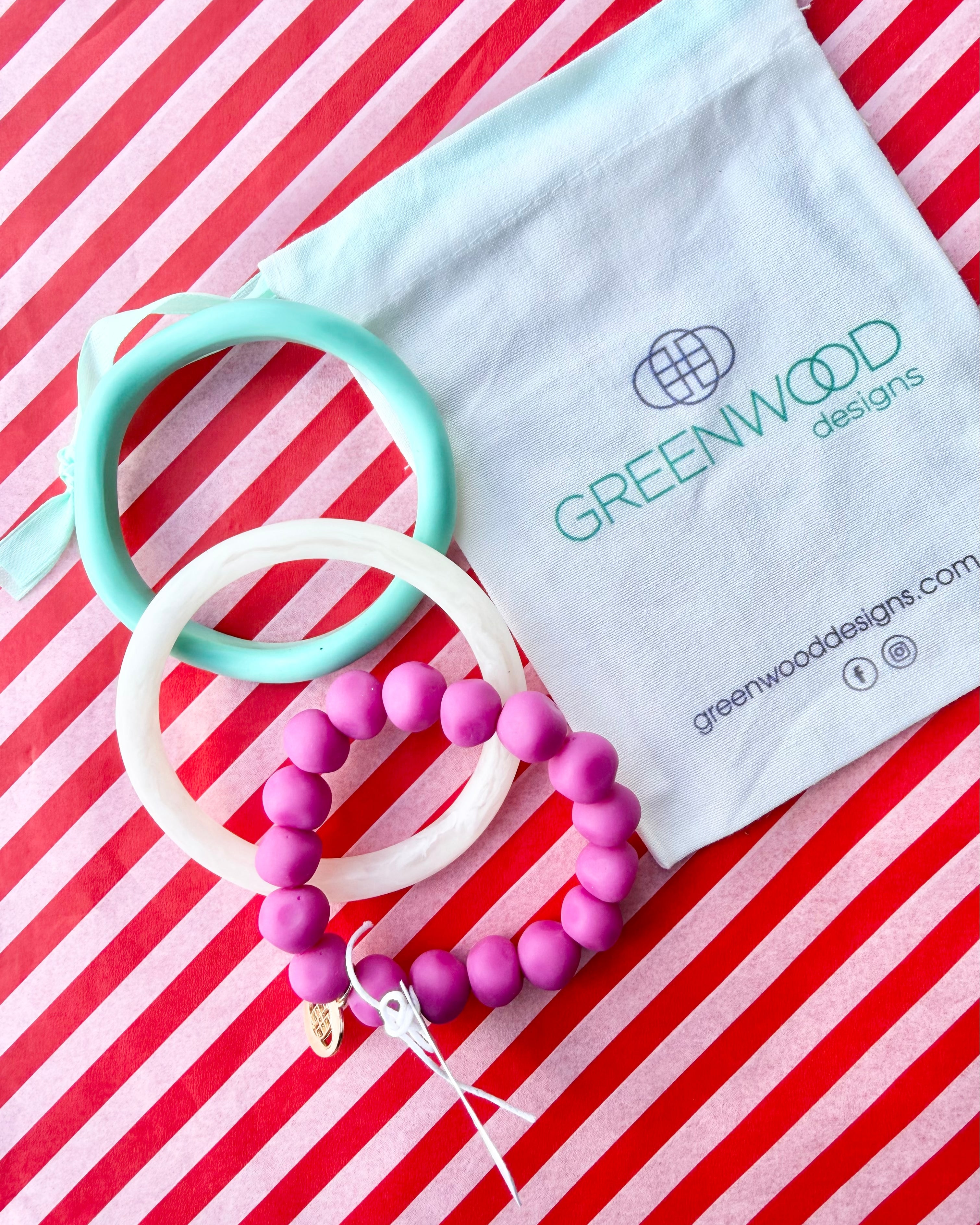 An aqua bangle, a white marbled bangle and a fuchsia rock bracelet laid on red and white striped paper beside a Greenwood Designs calico gift bag.
