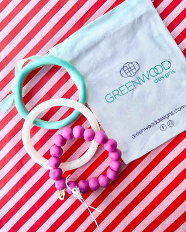 An aqua bangle, a white marbled bangle and a fuchsia rock bracelet laid on red and white striped paper beside a Greenwood Designs calico gift bag.
