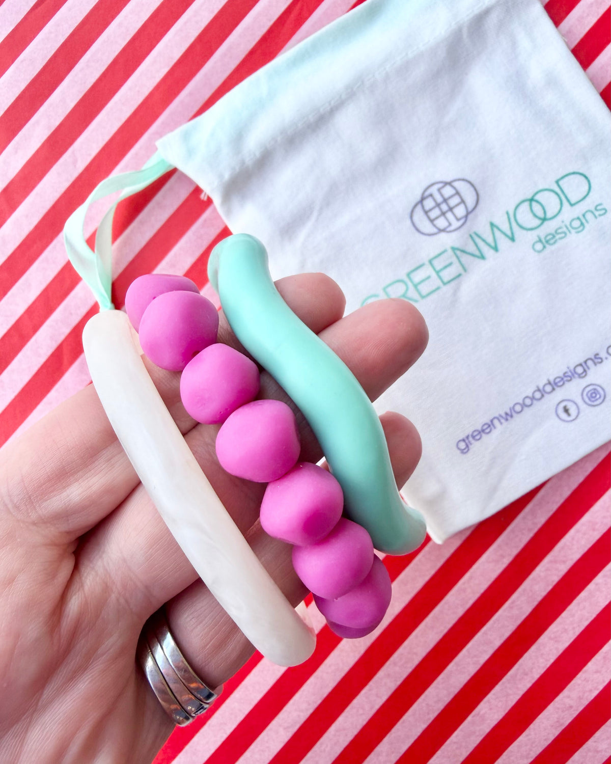 Hand holding a white marbled resin bangle, a fuchsia rock bracelet and an aqua wave bangle, with a Greenwood Designs calico gift bag on red and white striped paper.